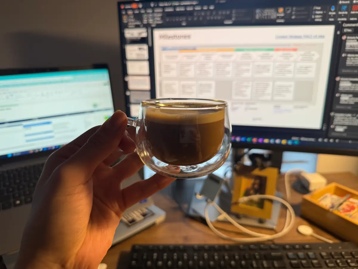 An espresso held up in front of a desk with a laptop and a monitor showing a project plan